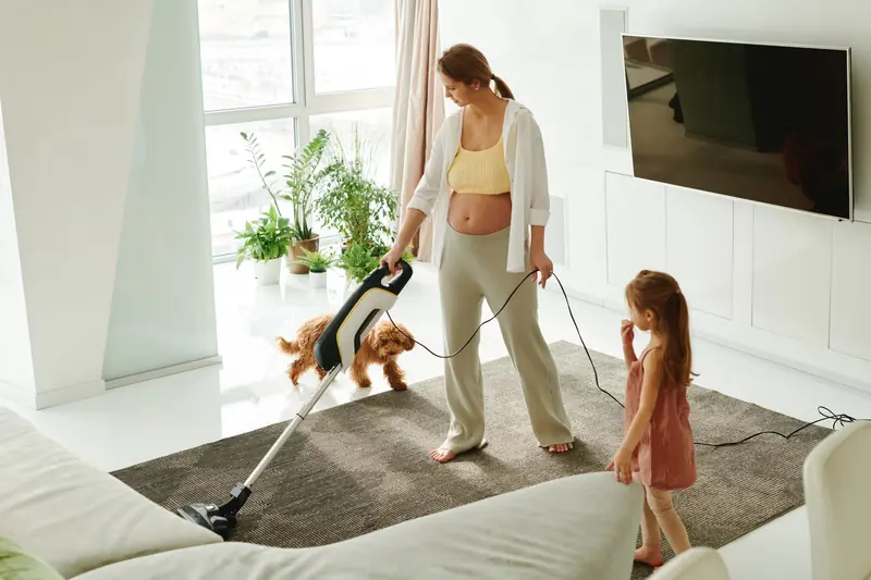 Pregnant mother cleaning carpet with vacuum as daughter and dog play indoors